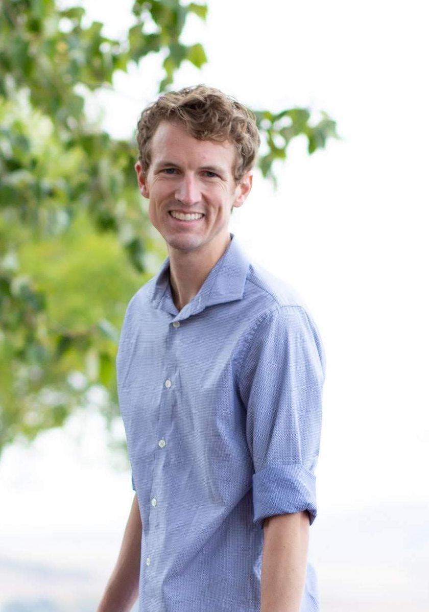Dr. Tim Johnson is standing on the waters edge in front of a tree with bright green leaves. He is wearing a blue button up shirt.