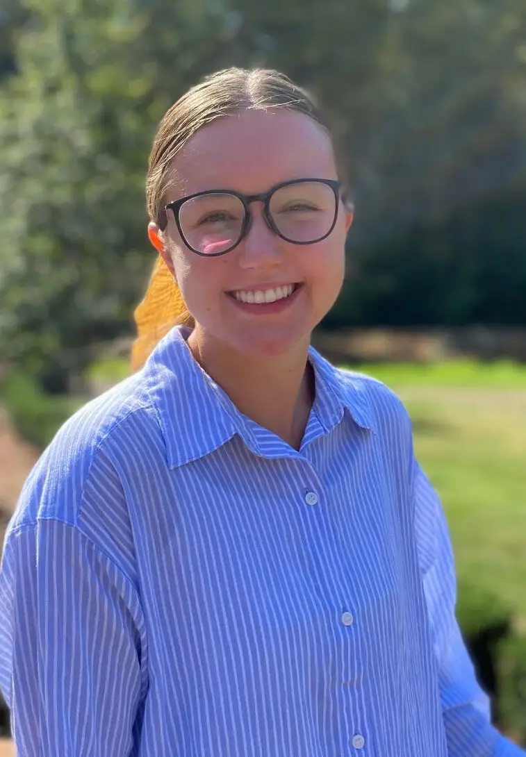Woman standing in the sunshine with her classes and a blue button up shirt.